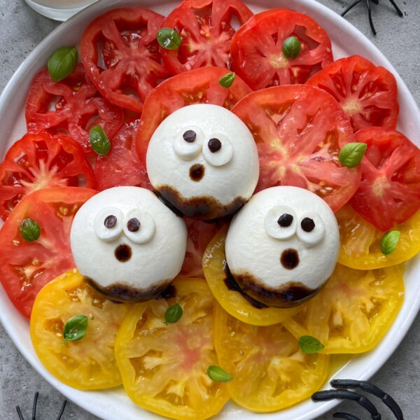 A plate with red and yellow tomato slices topped with mozzarella balls decorated as ghosts using balsamic glaze for eyes and mouths, garnished with small basil leaves—these playful bites make for fun, healthy food recipes.