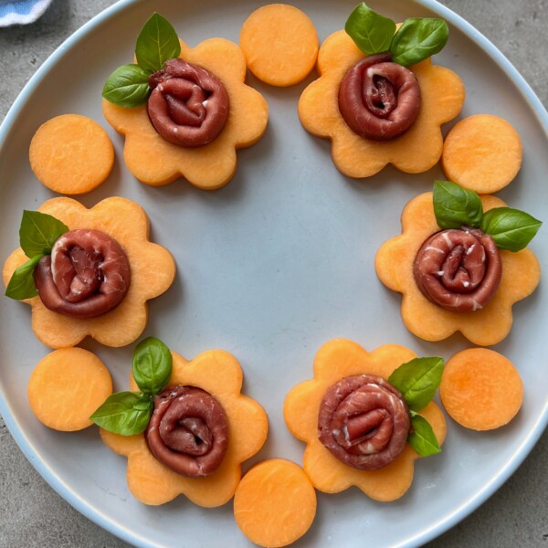 A round plate with six flower-shaped cantaloupe bites, each topped with rolled prosciutto and a basil leaf, arranged in a circle. Two round cantaloupe pieces complete this health-inspired appetizer.