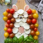 A creative food arrangement features mozzarella slices, cherry tomatoes, and basil forming a lamb face and wool on a wooden board—perfect for bites or healthy dishes. Basil leaves mimic grass, while red and yellow tomatoes add vibrant color.