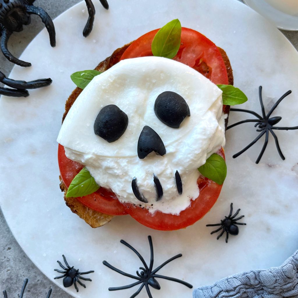 A creative food recipe: toast topped with tomato slices, mozzarella shaped like a skull, black olives for eyes and mouth, and basil leaves. Spooky bites are surrounded by plastic black spiders on a marble surface.