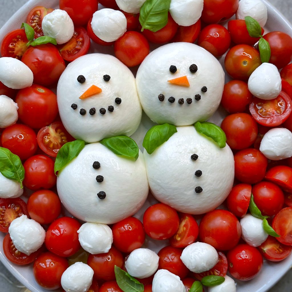 A plate with two snowmen made from mozzarella balls—perfect healthy bites—decorated with peppercorns, carrot slices, basil scarves, and surrounded by cherry tomatoes and more mozzarella balls for festive food recipes.