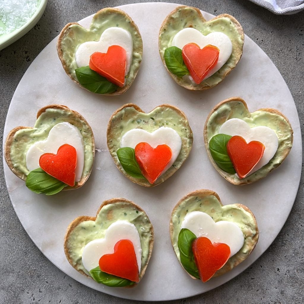 Seven heart-shaped Caprese Bites are arranged on a round white platter.
