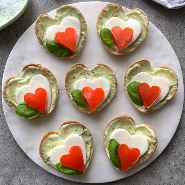 Seven heart-shaped Caprese Bites are arranged on a round white platter, each topped with creamy spread, basil, a heart-shaped mozzarella slice, and a heart-shaped tomato. Three feature basil beneath the cheese; four do not.