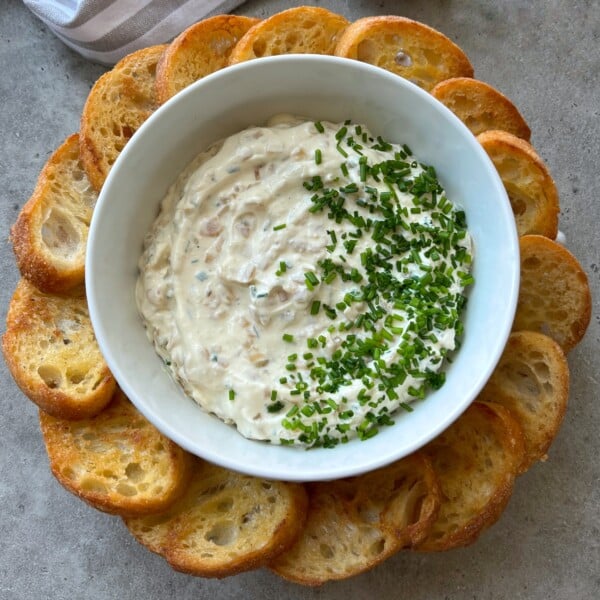 A white bowl filled with creamy caramelized onion dip topped with chopped chives is surrounded by a ring of toasted baguette slices on a gray surface.