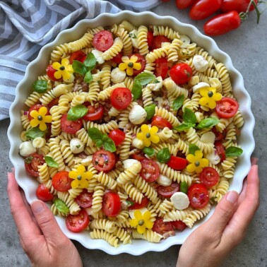 A round dish of rotini pasta salad with cherry tomatoes, mozzarella balls, basil leaves, and yellow bell pepper flowers, held by two hands. A striped cloth and tomatoes are in the background.