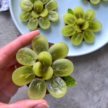 A hand holds a flower-shaped arrangement of sliced green grapes. More grape flowers are on a plate in the background, all neatly arranged in petal-like patterns.