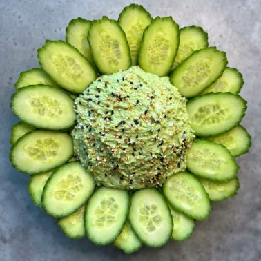 A round mound of green dip, topped with seasoning, sits on a gray surface. Slices of cucumber are arranged in a circular pattern around the dip, resembling flower petals.