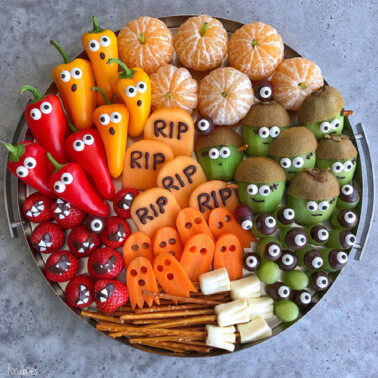 A tray of Halloween-themed fruits and veggies, including clementines, strawberries, peppers, kiwi, carrots, grapes, and cheese, decorated with candy eyes and “RIP” signs to resemble ghosts, monsters, and gravestones.
