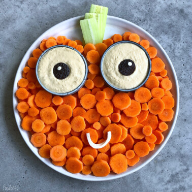 A round plate with carrot slices arranged as a face, two cups of hummus with olive pupils as eyes, celery sticks as hair, and a curved cheese slice as a smiling mouth on a gray surface.