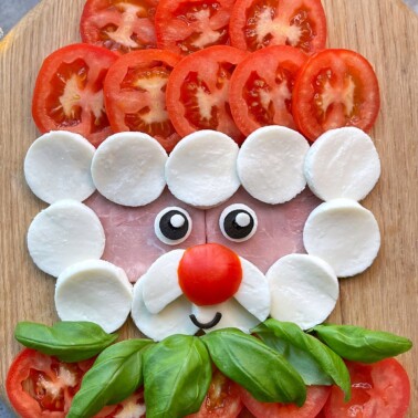 A face made from food: sliced tomatoes for hair, mozzarella rounds for cheeks and beard, a tomato nose, basil leaves as a mustache, ham as skin, and cartoon eyes on a wooden surface.