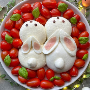 A plate of fresh mozzarella shaped like two bunnies, decorated with basil leaves, surrounded by cherry tomatoes. The bunnies have ears made from mozzarella and ham, with chocolate chips for eyes and noses.