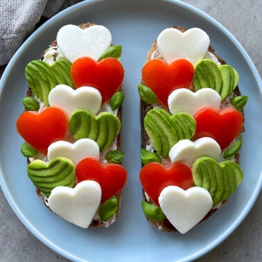 Two slices of bread topped with heart-shaped pieces of mozzarella, tomato, and avocado, garnished with fresh basil leaves, arranged neatly on a round plate.