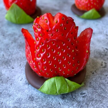 A strawberry carved to resemble a flower sits on a round chocolate base with green leaves, displayed on a gray surface. Other similar strawberry flowers are blurred in the background.