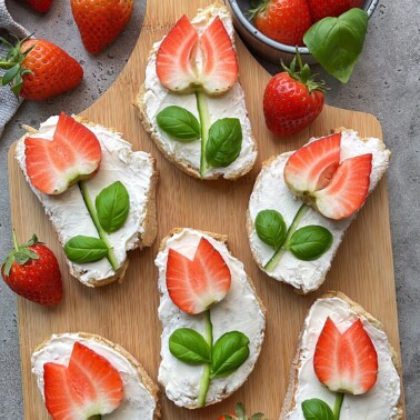 Slices of bread with white spread are topped with strawberry pieces and basil leaves, arranged to resemble tulip flowers. Whole strawberries and basil leaves are scattered on a wooden board and nearby surface.