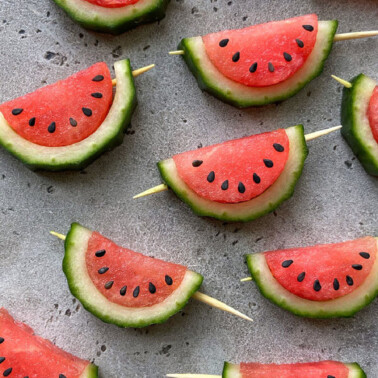 Slices of watermelon with rind and seeds are arranged on a gray surface, some served on wooden skewers.