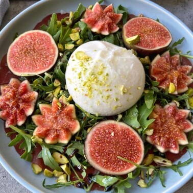 A plate of burrata cheese on a bed of arugula, surrounded by halved fresh figs with decorative cuts, and sprinkled with chopped pistachios.