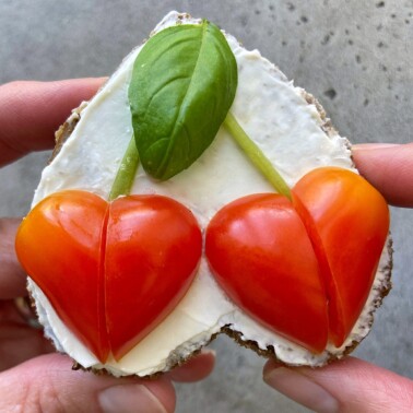 A heart-shaped piece of bread with cream cheese, topped with two red cherry tomato halves forming heart shapes and a green basil leaf. Two hands hold the bread against a gray background.