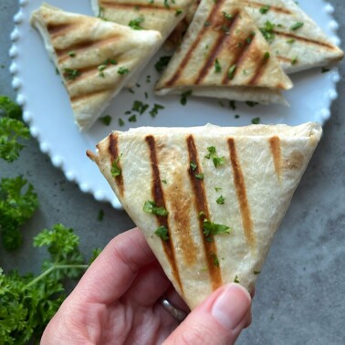 A hand holds a grilled tortilla triangle garnished with chopped herbs, with more tortilla triangles on a white plate in the background and fresh parsley nearby.