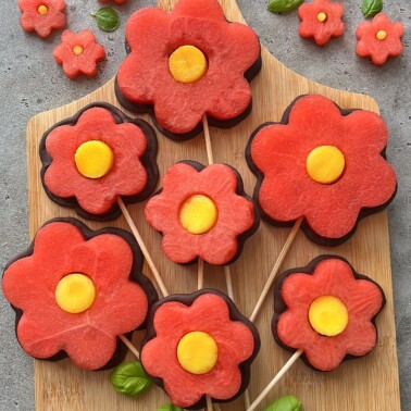 Flower-shaped watermelon slices with yellow centers are arranged on wooden skewers like lollipops, displayed on a wooden board with a few green basil leaves for decoration.