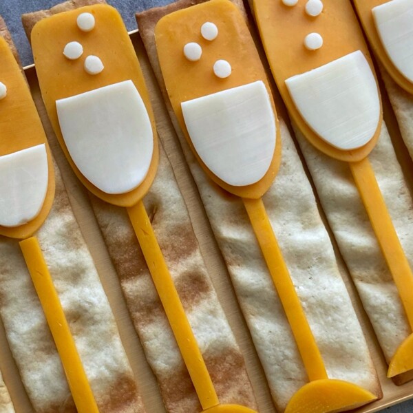 Rectangular crackers topped with cheese and white cheese slices arranged like champagne glasses, with small cheese circles as bubbles—these fun bites are perfect for party food recipes or festive dishes.