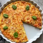 A Cheddar and Chicken Quiche with a golden, cheesy top and a slice removed, garnished with five fresh basil leaves, sits in a round white ceramic dish on a gray surface.