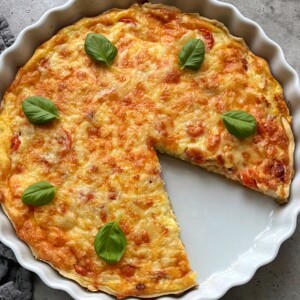 A Cheddar and Chicken Quiche with a golden, cheesy top and a slice removed, garnished with five fresh basil leaves, sits in a round white ceramic dish on a gray surface.