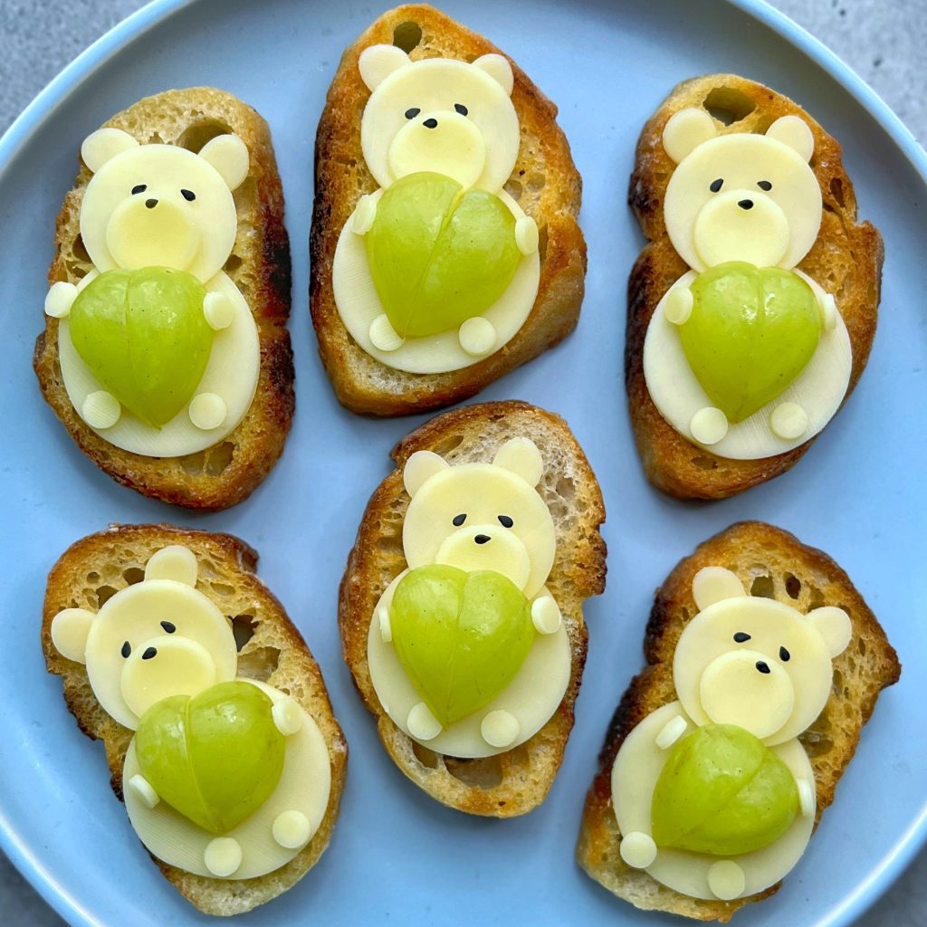 Six slices of toasted bread, each topped with cheese shaped like a bear holding a green grape “heart,” arranged on a round white plate. 