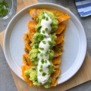 A plate of enchiladas topped with melted cheese, guacamole, sour cream, and chopped green onions is arranged neatly on a white plate over a wooden board—perfect for those seeking flavorful food recipes or delicious bites.