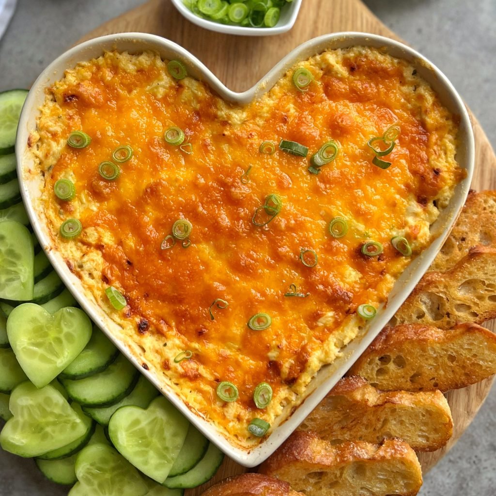 A heart-shaped dish filled with Cheesy Chicken Dip, topped with chopped green onions, surrounded by cucumber slices and toasted bread on a wooden board.
