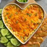 A heart-shaped dish filled with Cheesy Chicken Dip, topped with chopped green onions, surrounded by cucumber slices and toasted bread on a wooden board.
