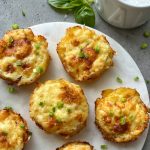 Six golden-brown baked egg muffins topped with chopped green herbs are arranged on a white round serving board, next to a sprig of basil and a bowl of cheesy smashed potatoes on a gray surface.