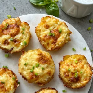 Six golden-brown baked egg muffins topped with chopped green herbs are arranged on a white round serving board, next to a sprig of basil and a bowl of cheesy smashed potatoes on a gray surface.