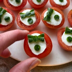 Close-up of a hand picking up a Cherry Tomato Appetizer topped with mozzarella, green bowtie-shaped herbs, and a small green dot. Several similar appetizers are arranged on a white plate.