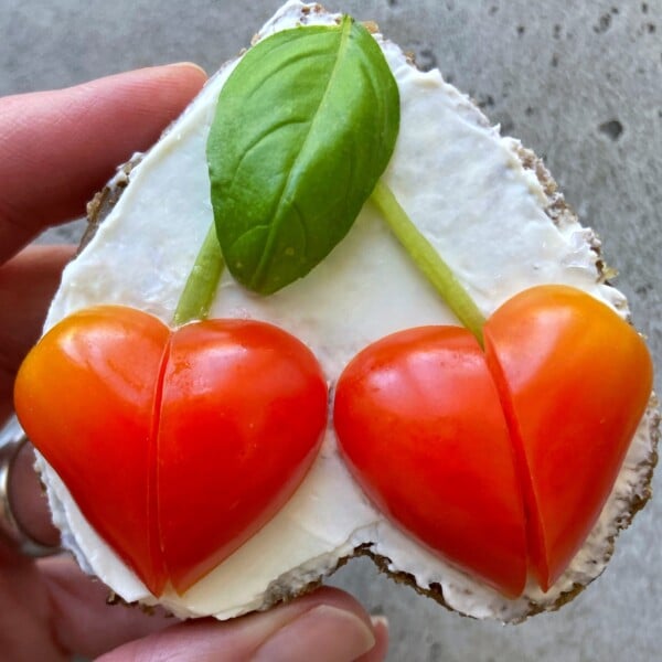 A hand holds a heart-shaped piece of bread topped with white spread, two cherry tomato halves arranged as hearts with green stems, and a basil leaf above them, resembling cherries—a delightful bite perfect for healthy food recipes.
