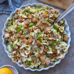 A large, round dish filled with Chicken Caesar Pasta Salad, featuring multicolored pasta, chopped lettuce, croutons, fresh basil leaves, and creamy dressing. Serving utensils rest in the bowl as a lemon sits nearby.