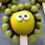 A yellow apple slice with candy eyes and green grapes arranged around it to look like a cartoon lion face on a popsicle stick—this playful bite makes healthy food fun for kids, perfect for creative recipes or snack dishes. Set on a gray surface.