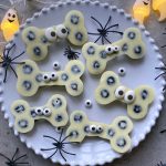 A white plate holds five bone-shaped bites with blueberries and candy eyes, surrounded by toy spiders and ghost string lights, creating a spooky, festive Halloween theme perfect for fun food recipes.