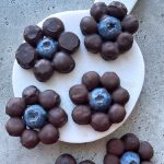 Five flower-shaped chocolate bites with a blueberry center are arranged on a white, round serving board atop a gray textured surface. Each treat is made from clustered dark chocolate-covered berries—perfect for health-conscious food lovers.