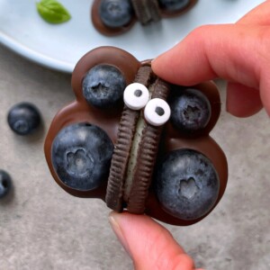 A hand holds a butterfly-shaped treat made from a chocolate-covered cookie with blueberries as wings and two small candy eyes on top—one of those creative food recipes that turns treats into playful bites. Other blueberries and a plate are in the background.