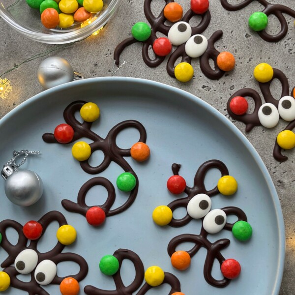 Chocolate squiggles decorated with colorful round candies and white candy eyes are arranged on a blue plate and gray surface, making these festive bites a fun addition to your food recipes collection, with a bowl of candies and silver ornaments nearby.