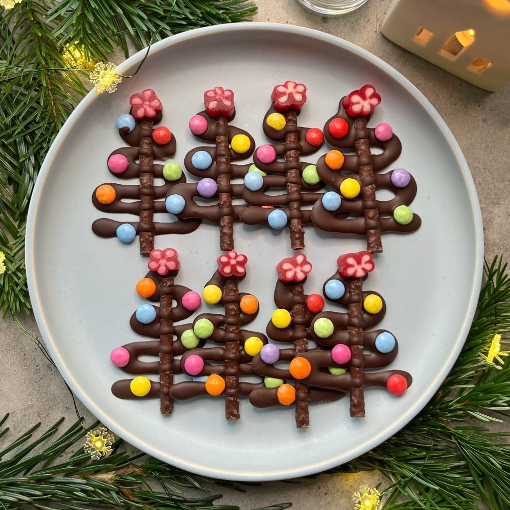 A plate with four chocolate Christmas tree bites, each decorated with colorful round candies and a red flower-shaped candy on top, surrounded by pine branches and string lights.