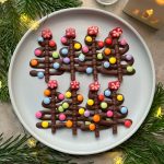 A plate with four chocolate Christmas tree bites, each decorated with colorful round candies and a red flower-shaped candy on top, surrounded by pine branches and string lights—perfect for festive food recipes.