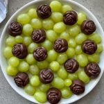 A white bowl filled with green grapes, topped with several chocolate candies shaped like roses, arranged evenly on the surface—perfect for health-conscious bites and creative food recipes.