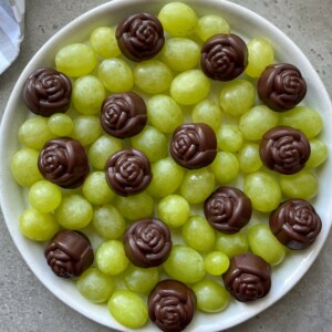 A white bowl filled with green grapes, topped with several chocolate candies shaped like roses, arranged evenly on the surface—perfect for health-conscious bites and creative food recipes.