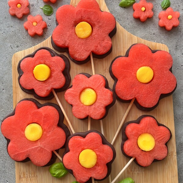 Sliced watermelon and yellow fruit arranged as flower shapes on sticks, resembling lollipops, displayed on a wooden board with small fruit flowers, basil leaves, and chocolate fruit skewers for a delightful touch.
