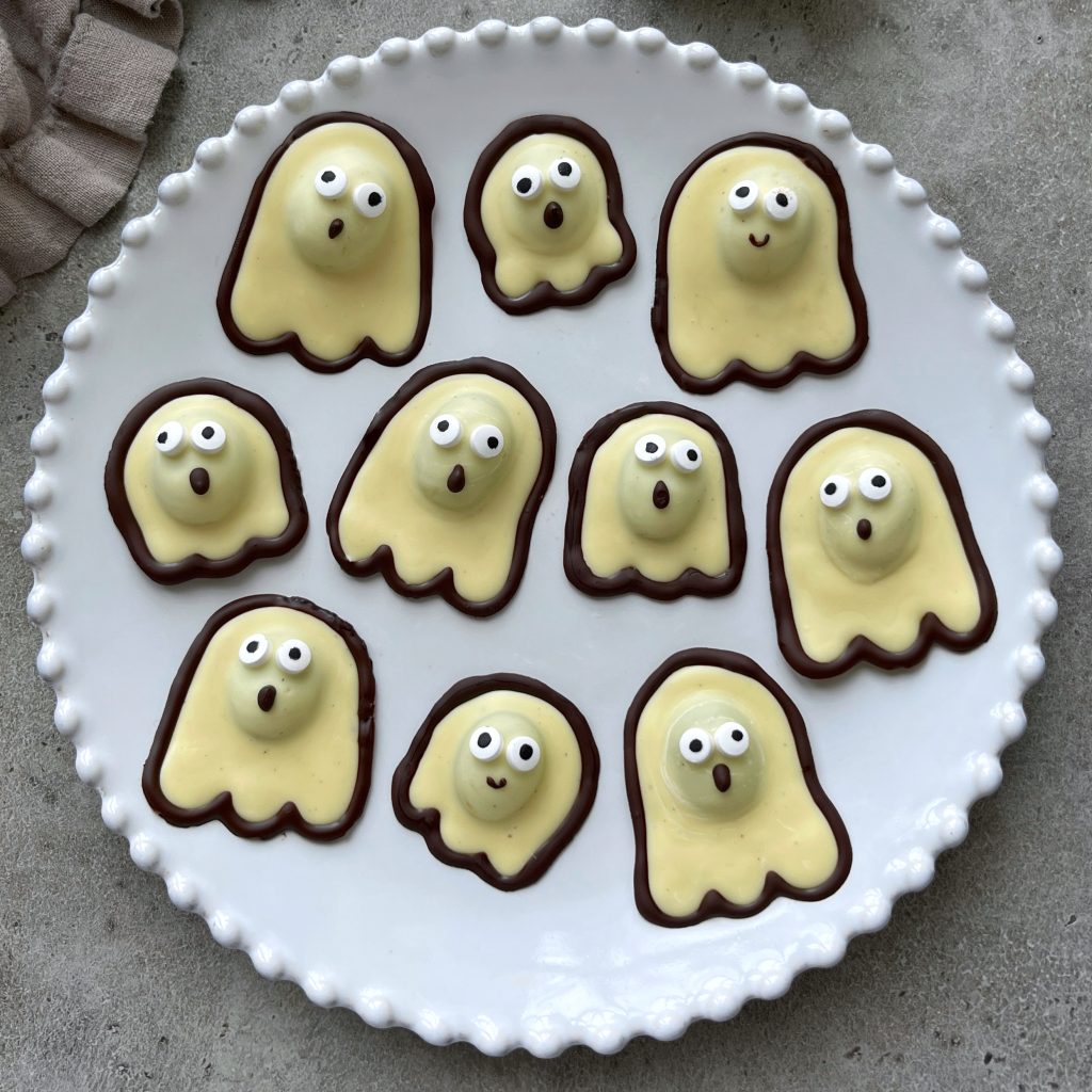 A white plate holds ten ghost-shaped cookies with yellow frosting, chocolate outlines, and icing faces, arranged on a gray surface—perfect bites for those who love fun food recipes.