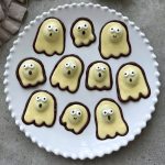 A white plate holds ten ghost-shaped cookies with yellow frosting, chocolate outlines, and icing faces, arranged on a gray surface—perfect bites for those who love fun food recipes.