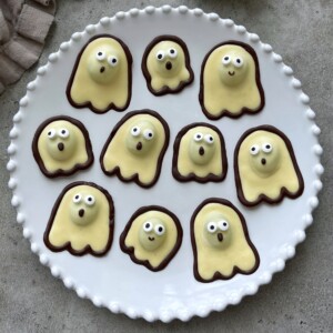 A white plate holds ten ghost-shaped cookies with yellow frosting, chocolate outlines, and icing faces, arranged on a gray surface—perfect bites for those who love fun food recipes.