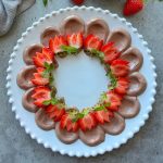 A round white plate with dollops of chocolate mousse and chocolate Greek yogurt dip piped in a circle, topped with halved strawberries and fresh mint leaves, forming a decorative ring on a gray surface.