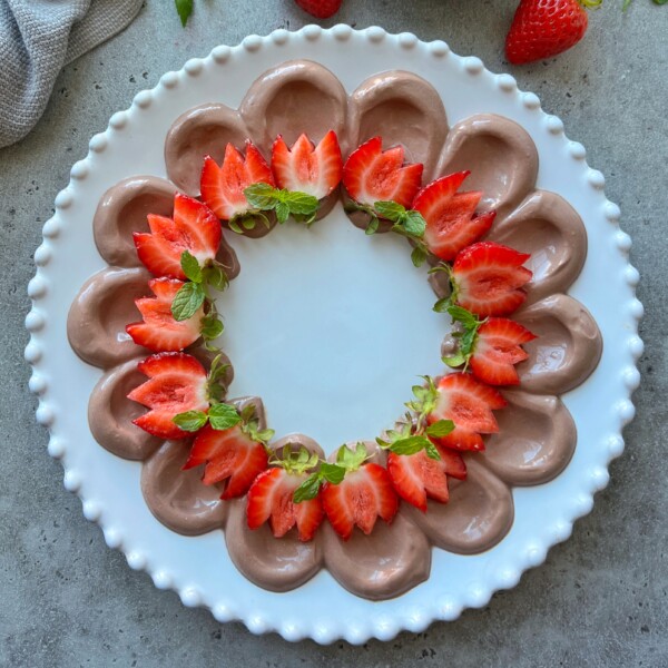 A round white plate with dollops of chocolate mousse and chocolate Greek yogurt dip piped in a circle, topped with halved strawberries and fresh mint leaves, forming a decorative ring on a gray surface.